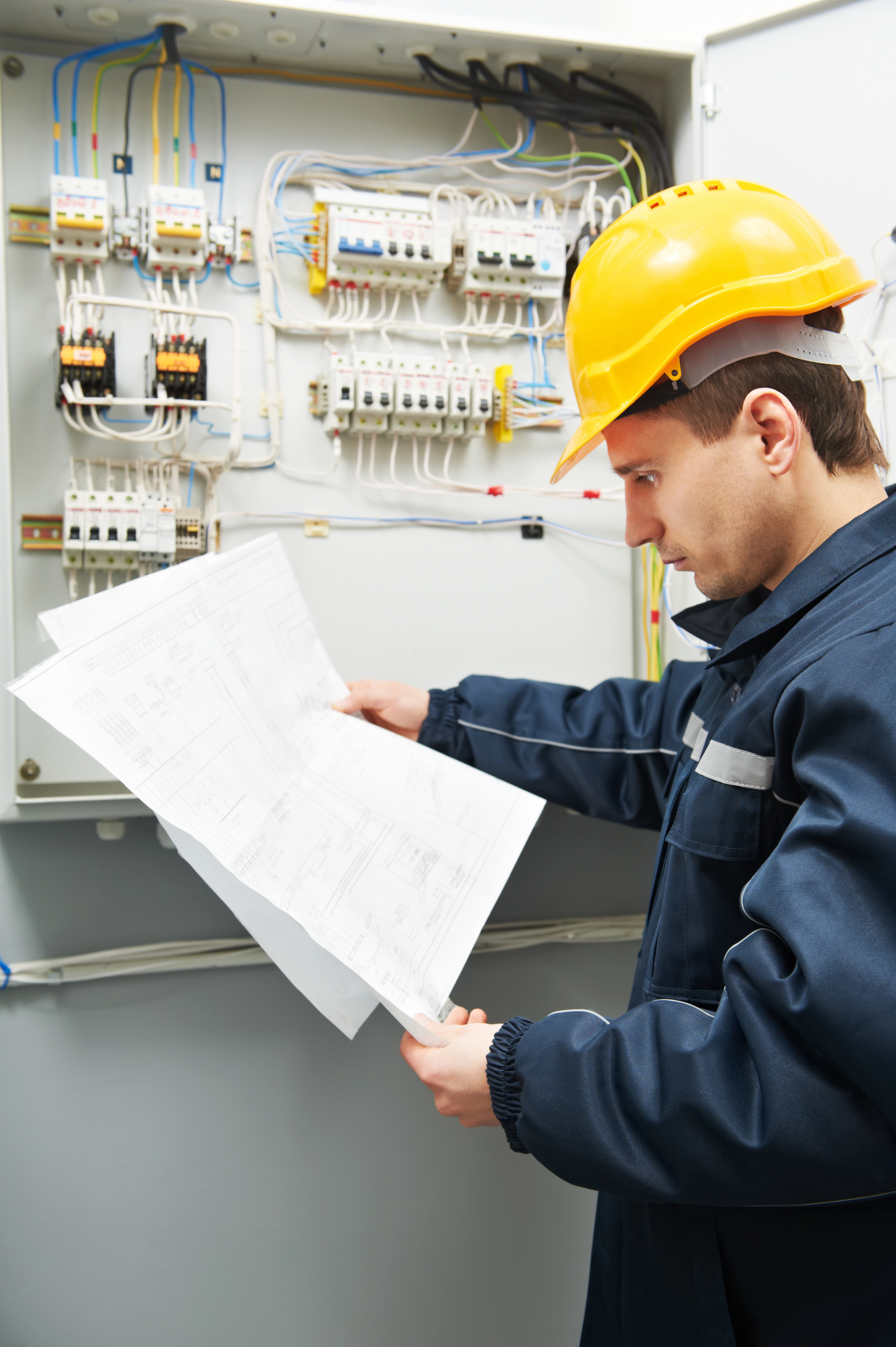 Engineer in jacket and yellow hard hat standing by an electrical board looking over paperwork
