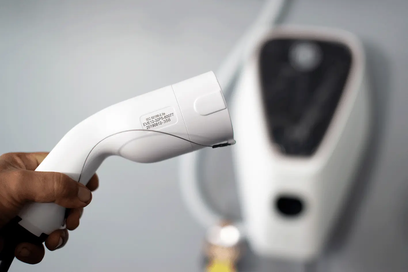 A hand holding a white electric vehicle charging plug in front of a charging point
